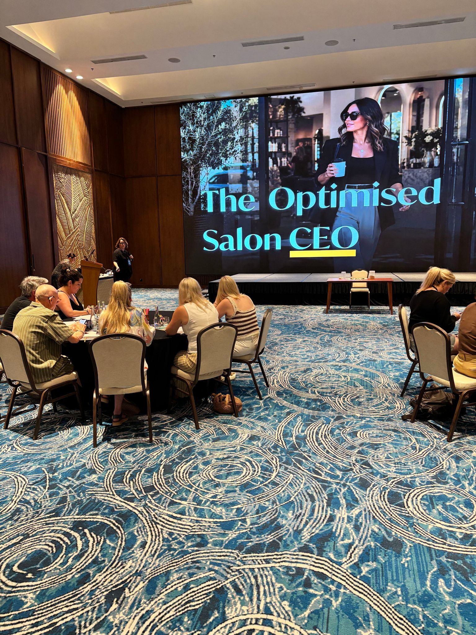 Salon owners seated at tables at a business event in front of a large presentation screen