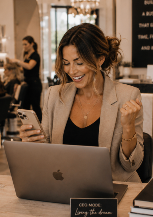 A smiling salon owner celebrating while looking at her phone and laptop inside her salon